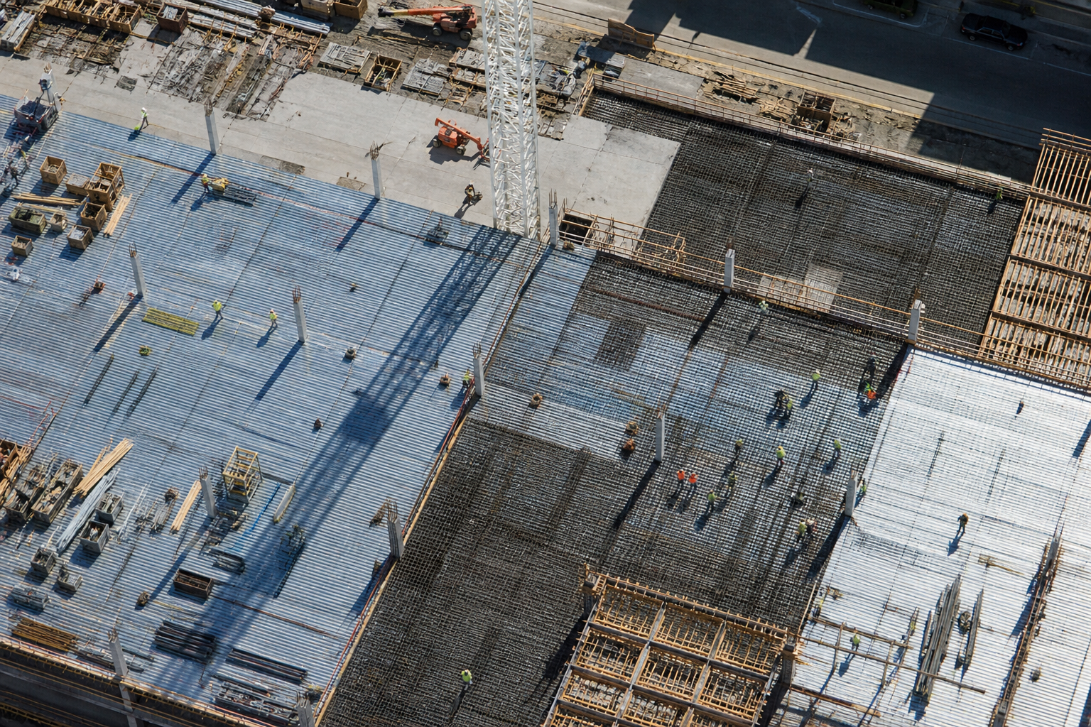 Overhead view of a large floor plate with rebar, decking, crane shadow, and trades on the deck.